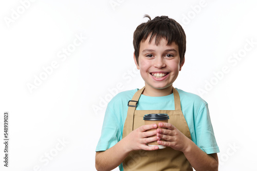 Multi-ethnic handsome preteen boy, wearing turquoise t-shirt and beige chef's apron holding an eco paper cup of takeaway coffee, smiling toothy smile looking at camera, isolated over white background