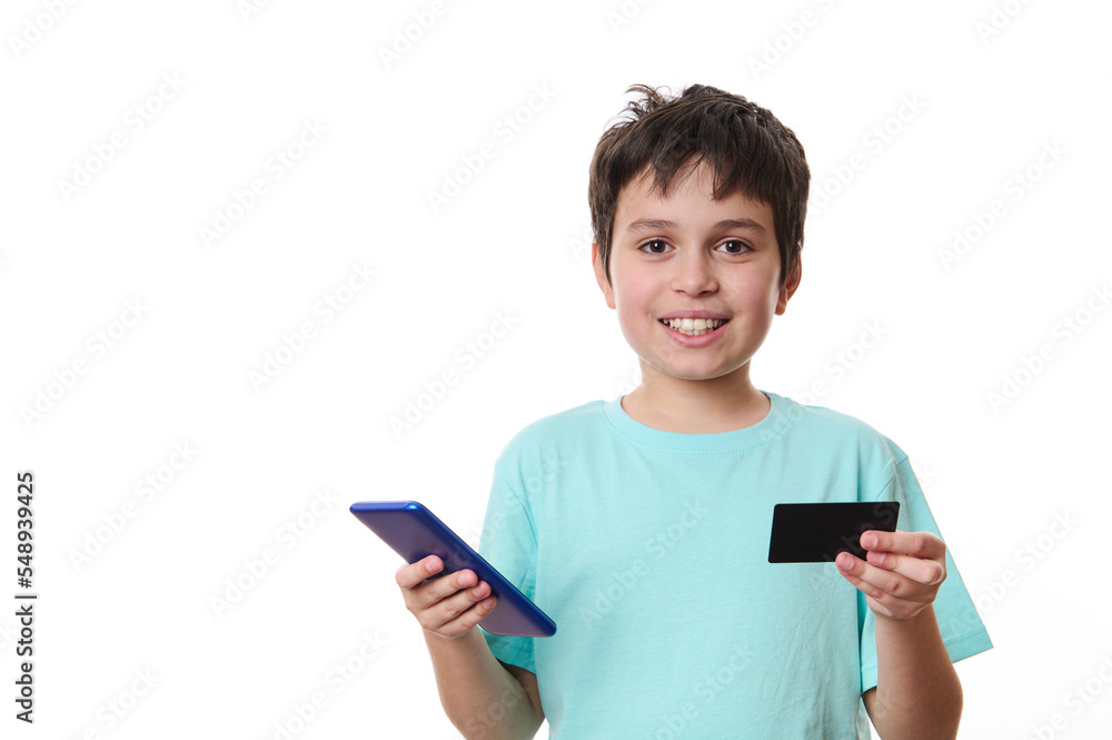Adorable dark-haired Caucasian preteen boy, smart school kid in blue t-shirt with smartphone and plastic credit card, isolated on white background. Internet banking. Cashless payment. Online shopping Adorable dark-haired Caucasian preteen boy, smart school kid in blue t-shirt with smartphone and plastic credit card, isolated on white background. Internet banking. Cashless payment. Online shopping