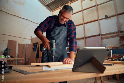 Mid adult man smiling while using drill on timber next to digital tablet in woodworking factory