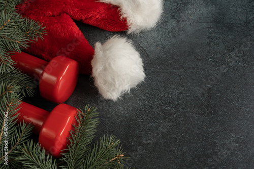 Two heavy dumbbells, red Santa Claus hat and Christmas tree branches. Healthy fitness lifestyle holiday season winter composition. Gym workout and sport training concept, with copy space.