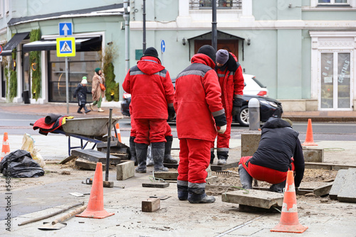 Road works, street repairs in city. Workers in red uniform lying stone blocks on sidewalk