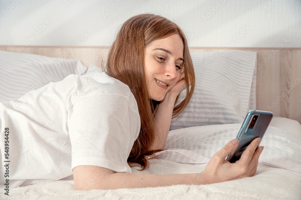 Woman with smartphone in bed at home or hotel in the morning. Lady relaxing in a nightgown. Girl wearing nightie on clean white bedding, cozy blanket Woman with smartphone in bed at home or hotel in the morning. Lady relaxing in a nightgown. Girl wearing nightie on clean white bedding, cozy blanket