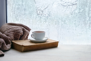 tea cup, book, sweater and winter frozen window. cozy mood, home comfort in snowy cold weather. festive winter season. Christmas, New Year holidays background. copy space