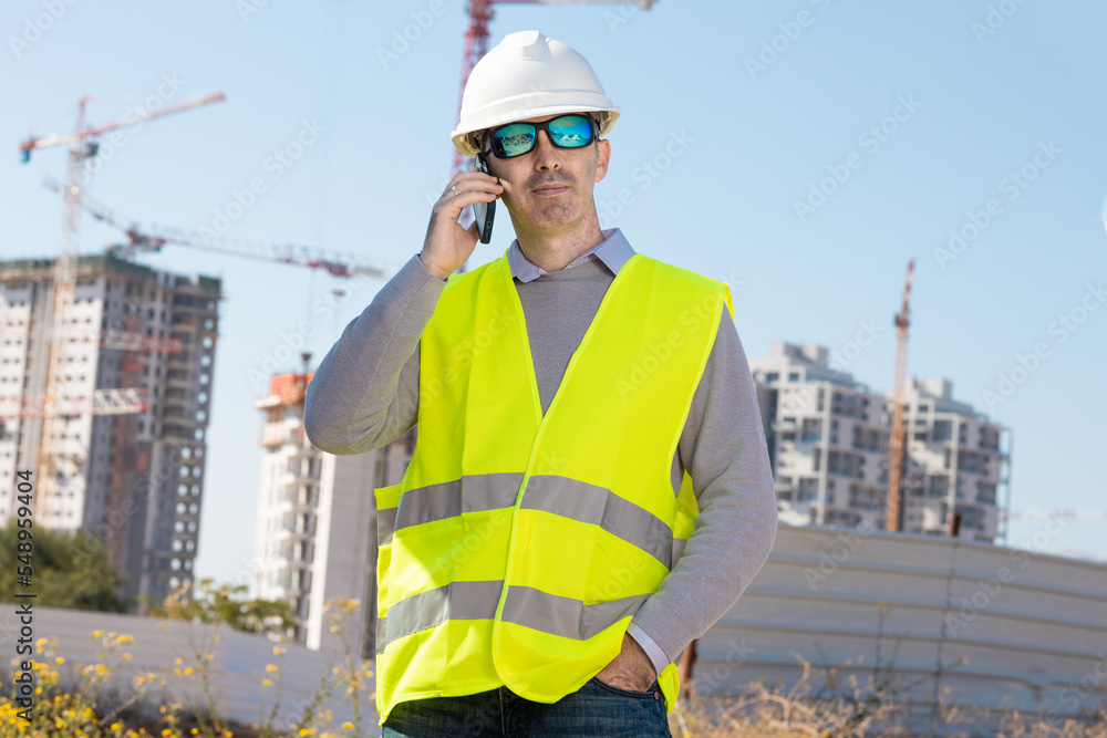 Professional builder standing talking on a phone in front of the construction site Professional builder standing talking on a phone in front of the construction site