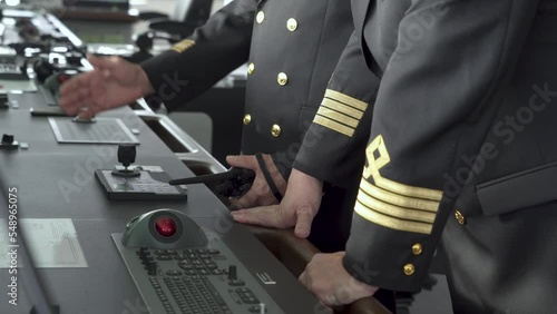 Ship's bridge. Officers stand at control panel. Hand turns joystick and hold radio. hief Mate, second mate patches on jackets. photo