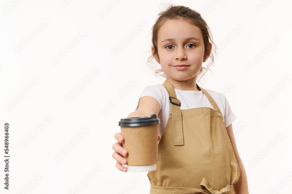 Caucasian lovely little girl wearing a beige chef's apron, handing out on camera a takeaway hot tea cofee drink in cardboard mug, isolated on white, copy space for text. Beverage in paper cup to go
