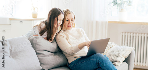 Mom and daughter spend time at home on the couch in the living room with a computer. Online communication.