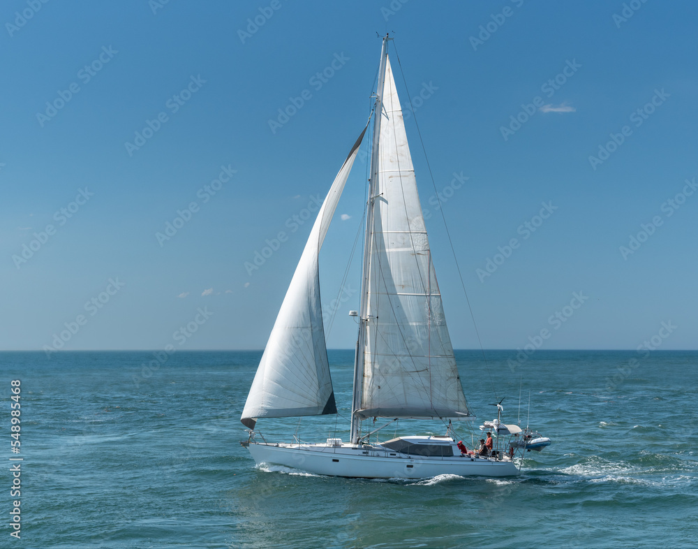 A white yacht under sail on the ocean with three unidentified people on board.