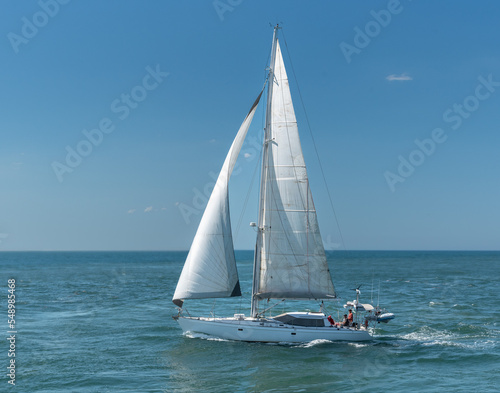 A white yacht under sail on the ocean with three unidentified people on board.