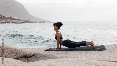 Woman exercising on a mat by ocean. Slim female doing stretching pose outdoors at sunset. © Artem Varnitsin