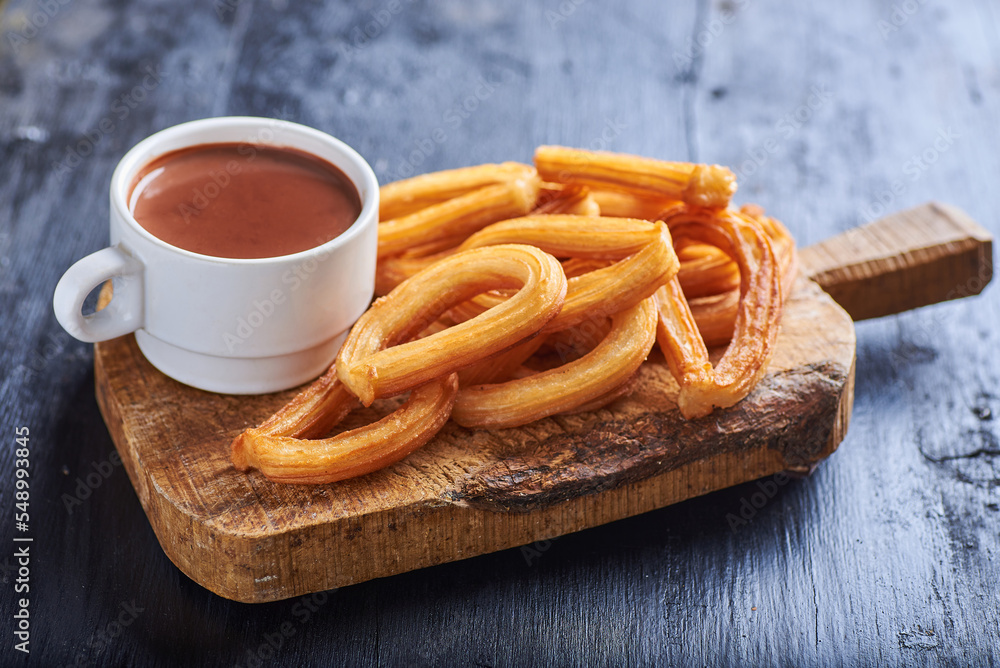 Chocolate en una taza de color blanco sobre una superficie rústica y rodeada de churros y un fondo negro Chocolate en una taza de color blanco sobre una superficie rústica y rodeada de churros y un fondo negro