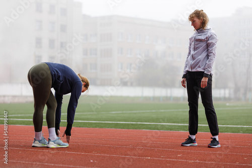 Two young women doing running training stretching legs on the track in the foggy morning. Coach and trainee at the athletics training