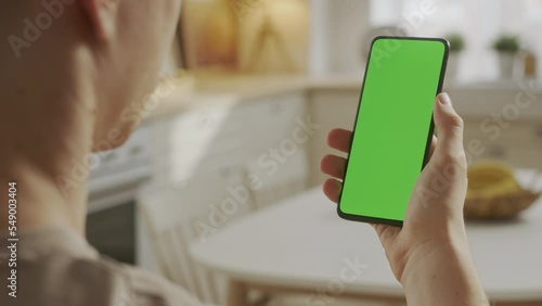 Back View of Young Man at Home Sitting on Kitchen Room With Green Mock-up Screen Smartphone. Boy is Watcing Content Without Touching Gadget Screen. Modent Technology photo