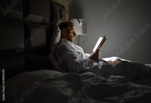 Canvas Print Midle-aged caucasian man relaxing in bed reading bestseller novel paper book with bedside lamp turned on