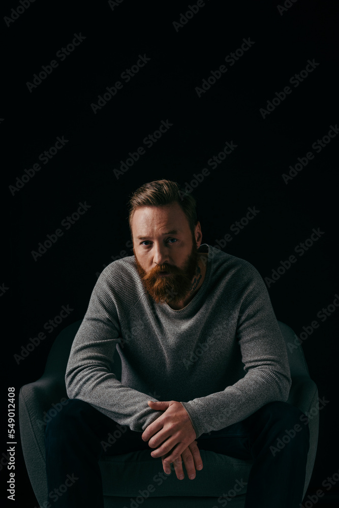 Fashionable man in jumper looking at camera near armchair isolated on black.