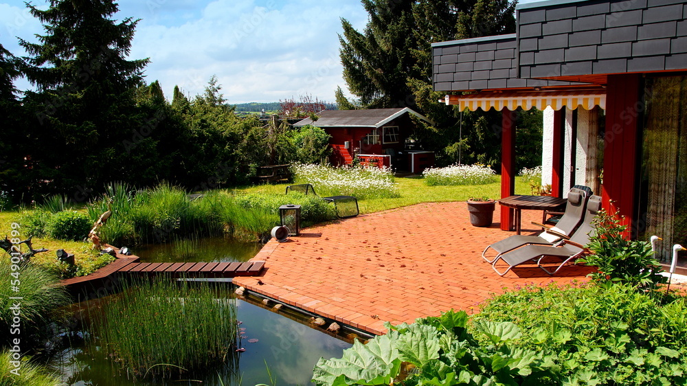 schönes Haus mit an roter Terrasse angrenzendem Teich mit Brücke in malerischem Garten bei blauem Himmel