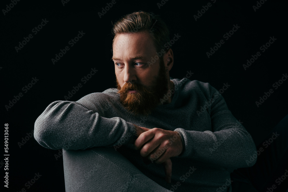 Bearded model in grey jumper looking away while sitting on armchair isolated on black.