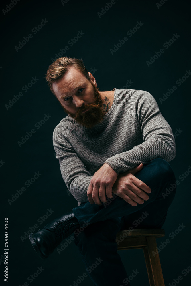 Tattooed and bearded model in jumper posing on chair isolated on black.