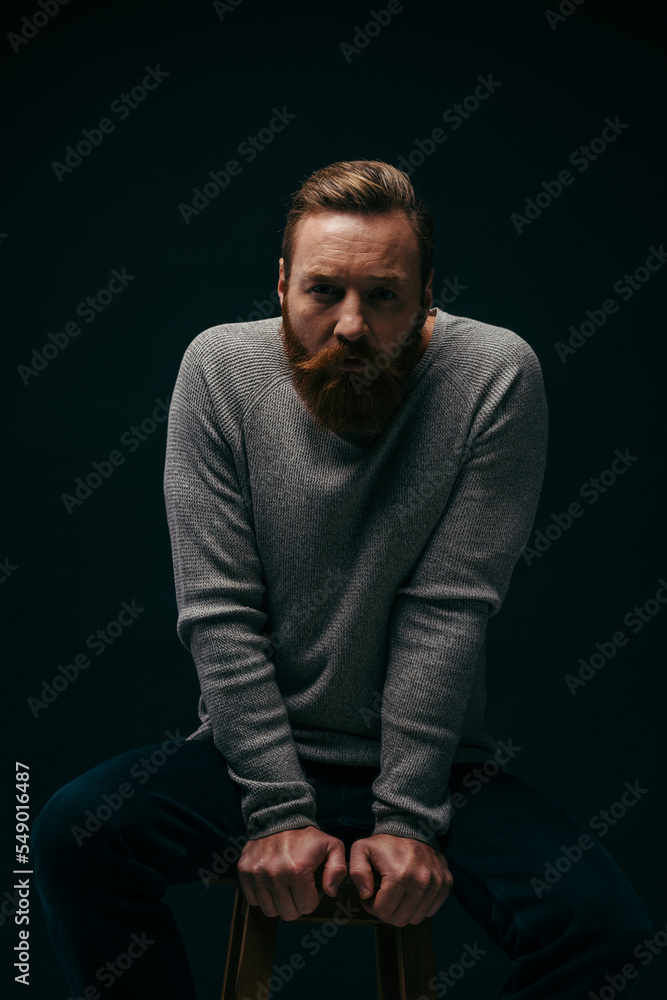 Portrait of bearded man posing while sitting on chair isolated on black with shadow.