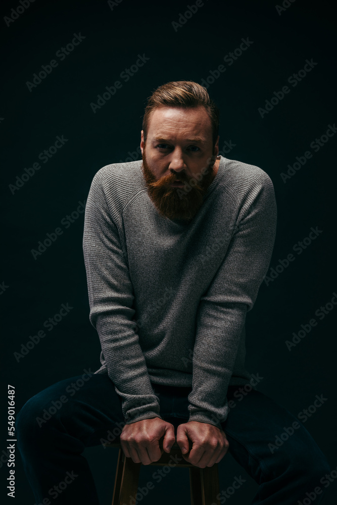 Fototapeta premium Portrait of bearded man posing while sitting on chair isolated on black with shadow.