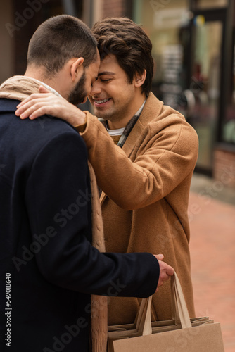 trendy gay man with closed eyes smiling near boyfriend holding shopping bags on urban street.