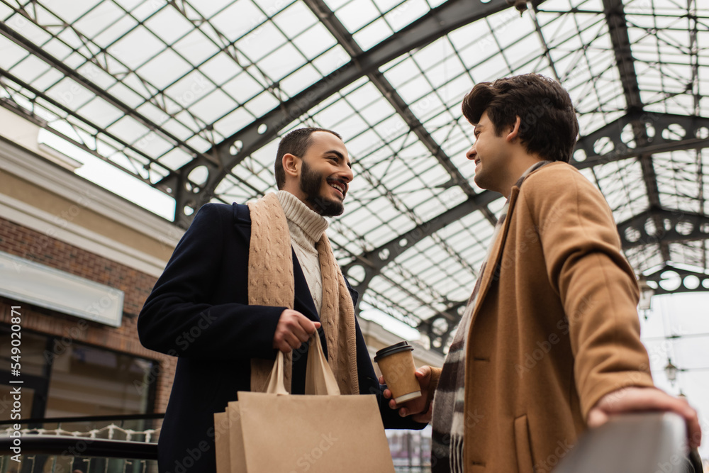 low angle view of stylish homosexual couple with shopping bags and paper cup under transparent roof outdoors. low angle view of stylish homosexual couple with shopping bags and paper cup under transparent roof outdoors.