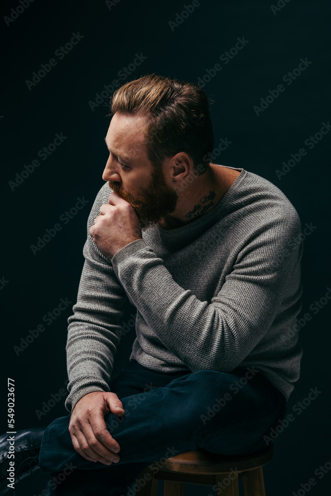 Side view of stylish tattooed man in grey jumper sitting on chair isolated on black. Side view of stylish tattooed man in grey jumper sitting on chair isolated on black.