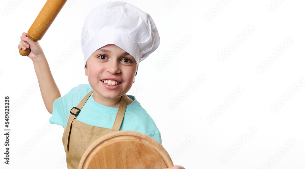 Close-up of a smiling mischievous boy - baker confectioner wearing chef's hat and apron, fighting with wooden board and rolling pin in his hands, isolated on white background. Bakery advertising shot. Close-up of a smiling mischievous boy - baker confectioner wearing chef's hat and apron, fighting with wooden board and rolling pin in his hands, isolated on white background. Bakery advertising shot.