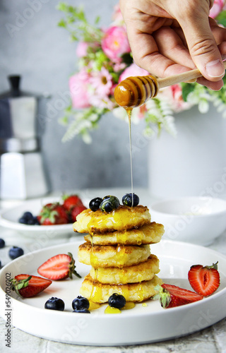 Cottage cheese pancakes with fresh strawberries, blueberries and honey on a white plate with a hand pooring honey. Tasty breakfast food. Homemade syrniki. Coffee time. Grey background photo