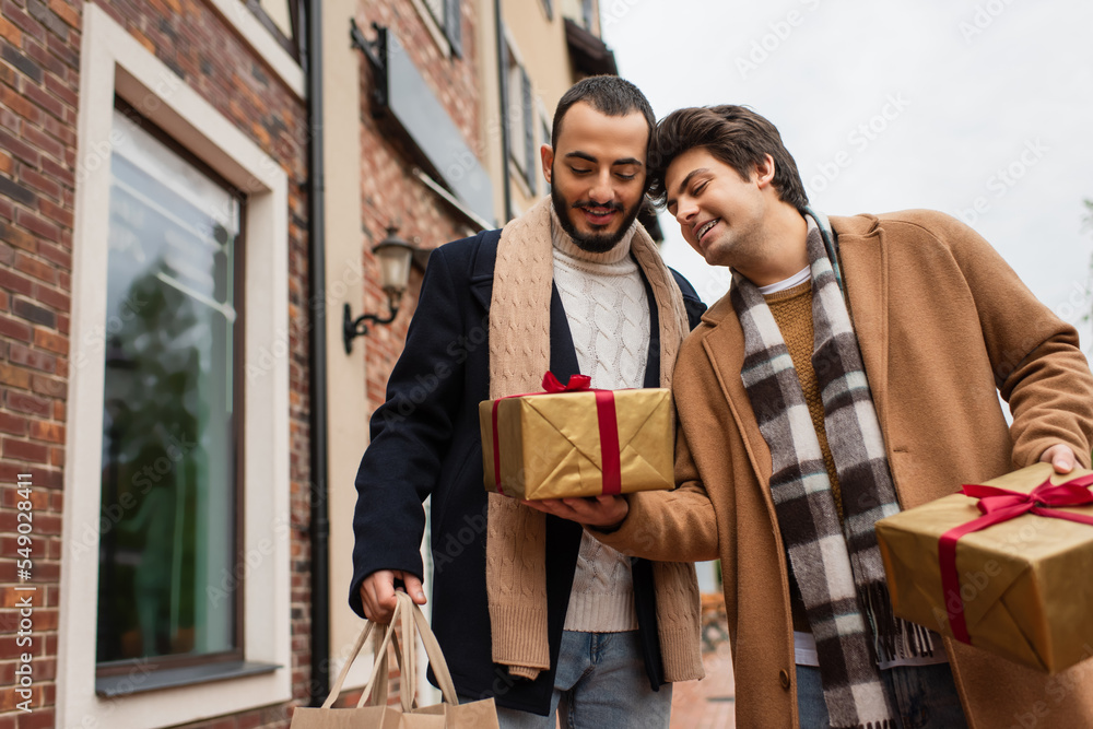 Obraz premium young gay man holding gift boxes near happy and trendy boyfriend with shopping bags on urban street.