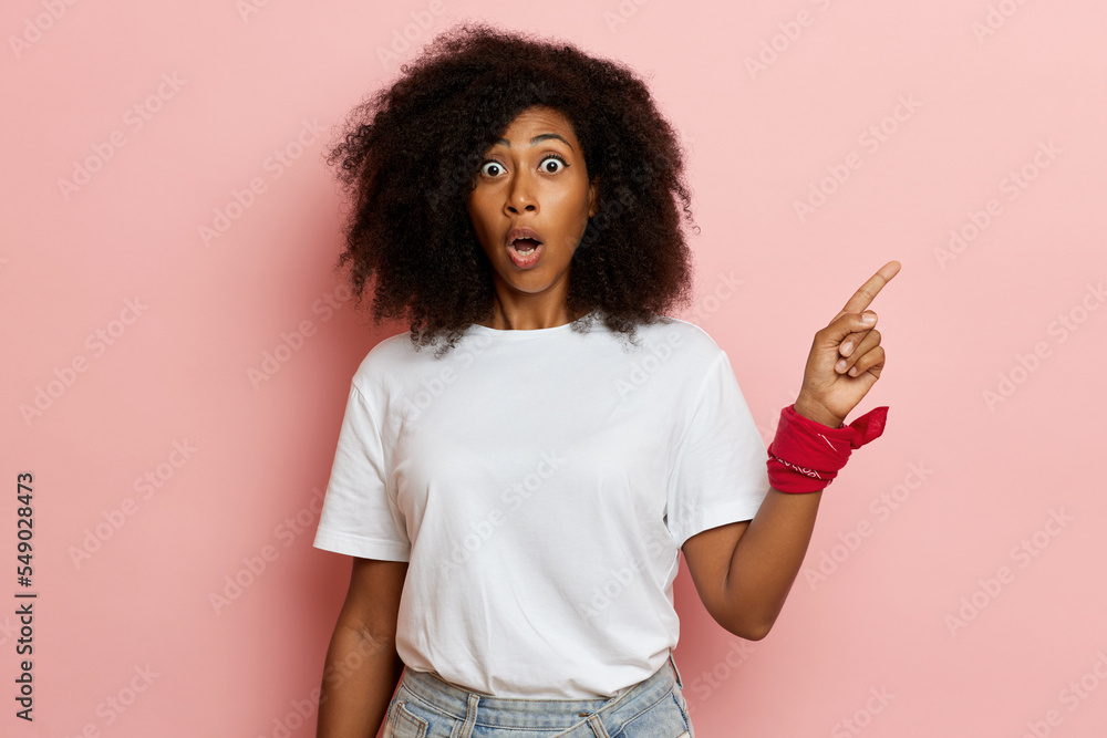 Young curly haired woman, shows finger a side to the left, open eyes and mouth widely, isolated over pink background Young curly haired woman, shows finger a side to the left, open eyes and mouth widely, isolated over pink background