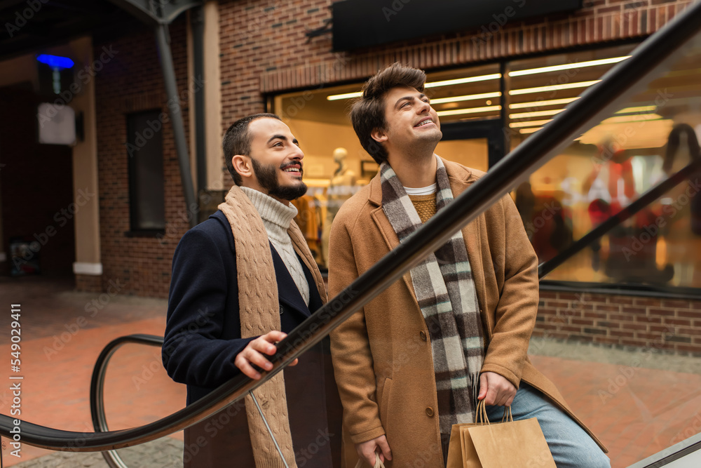 smiling bearded man with boyfriend holding shopping bags and looking away on escalator.