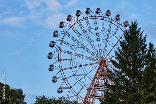 Big ferris wheel over background of blue sky with fluffy clouds and coniferous trees.
