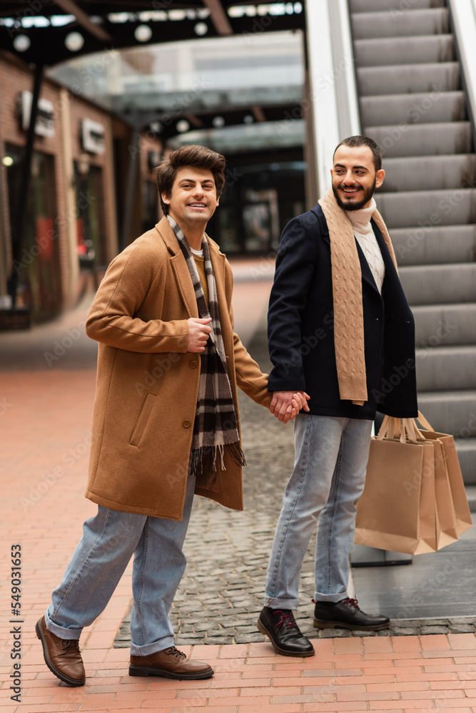 gay man in beige coat and plaid scarf holding hands with smiling bearded boyfriend with shopping bags near escalator.