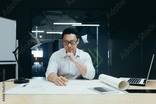 A handsome young man, an Asian architect, designer, engineer, sits at a desk in the office at a table with notebooks and documents. He concentrates on making drawings, developing a plan.