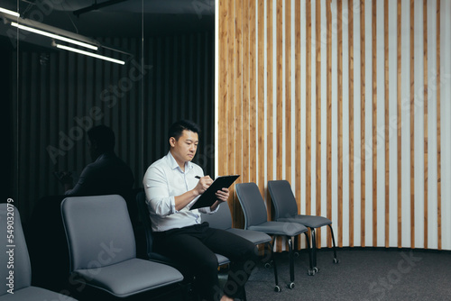 A young handsome Asian man is sitting seriously in the lobby of the office center, waiting for an interview, holding a folder with papers and documents in his hands. He is filling out a questionnaire.