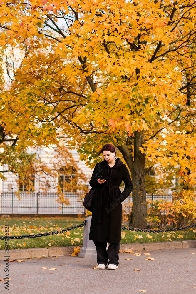 A young teenage girl in a black coat stands with her phone in her hand and looks into it against a backdrop of maple trees with yellow leaves in the fall. A young teenage girl in a black coat stands with her phone in her hand and looks into it against a backdrop of maple trees with yellow leaves in the fall.