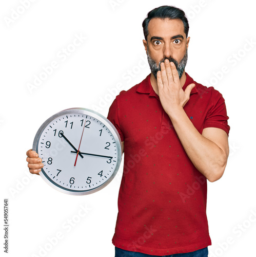 Middle aged man with beard holding big clock covering mouth with hand, shocked and afraid for mistake. surprised expression