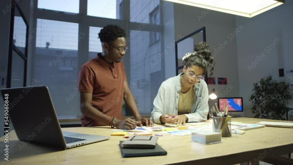 Zoom in shot of Latin businesswoman using sticky notes and talking to black colleague while working together on creative project late in office Zoom in shot of Latin businesswoman using sticky notes and talking to black colleague while working together on creative project late in office