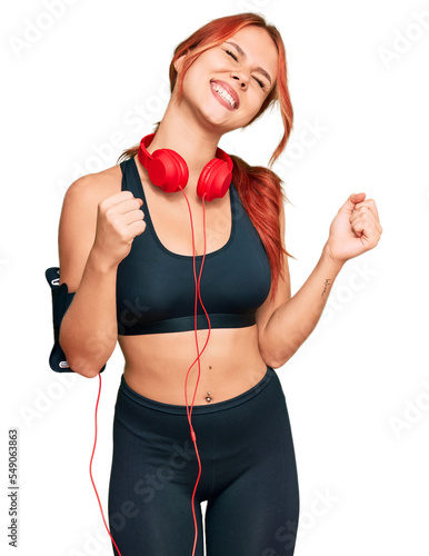 Young redhead woman wearing gym clothes and using headphones very happy and excited doing winner gesture with arms raised, smiling and screaming for success. celebration concept.