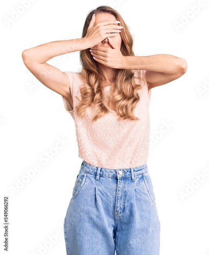 Young beautiful caucasian woman with blond hair wearing casual clothes covering eyes and mouth with hands, surprised and shocked. hiding emotion