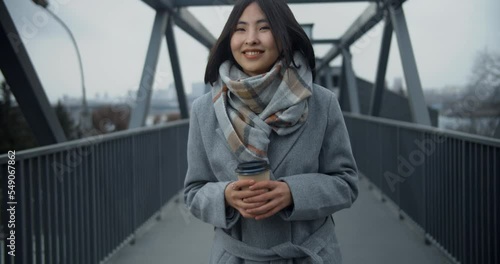 A young beautiful Asian woman in a coat with a cup of coffee walking along the transition in the middle of an empty cold city. Good mood, smiling. Attractive girl. Autumn. 