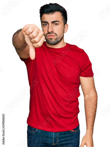 Hispanic man with beard wearing casual red t shirt looking unhappy and angry showing rejection and negative with thumbs down gesture. bad expression.