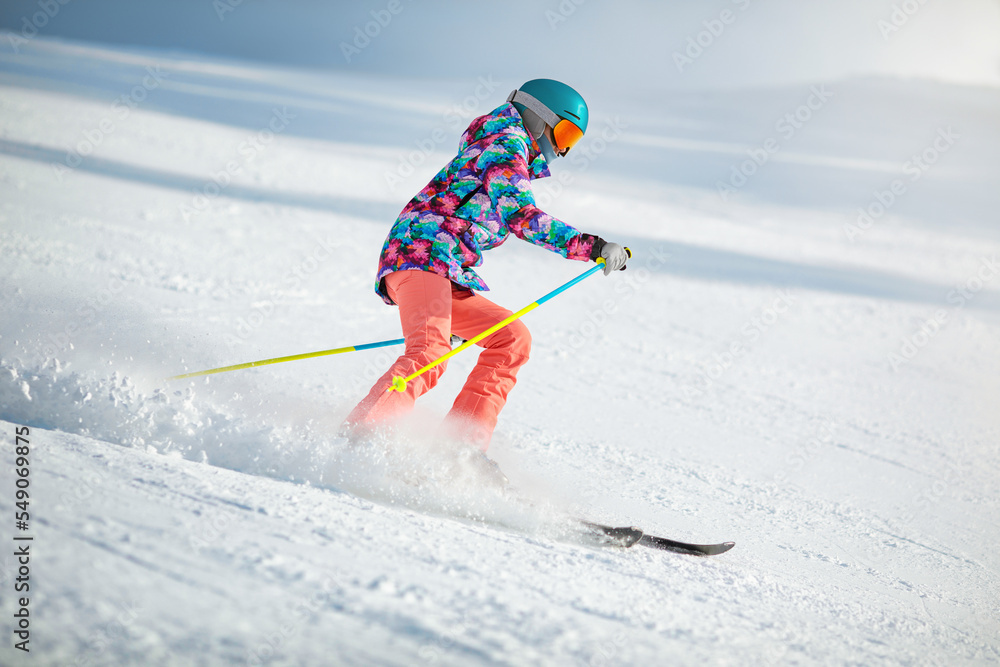 Young female skier in colourful clothes sliding down the slope on a sunny day at a mountain resort.