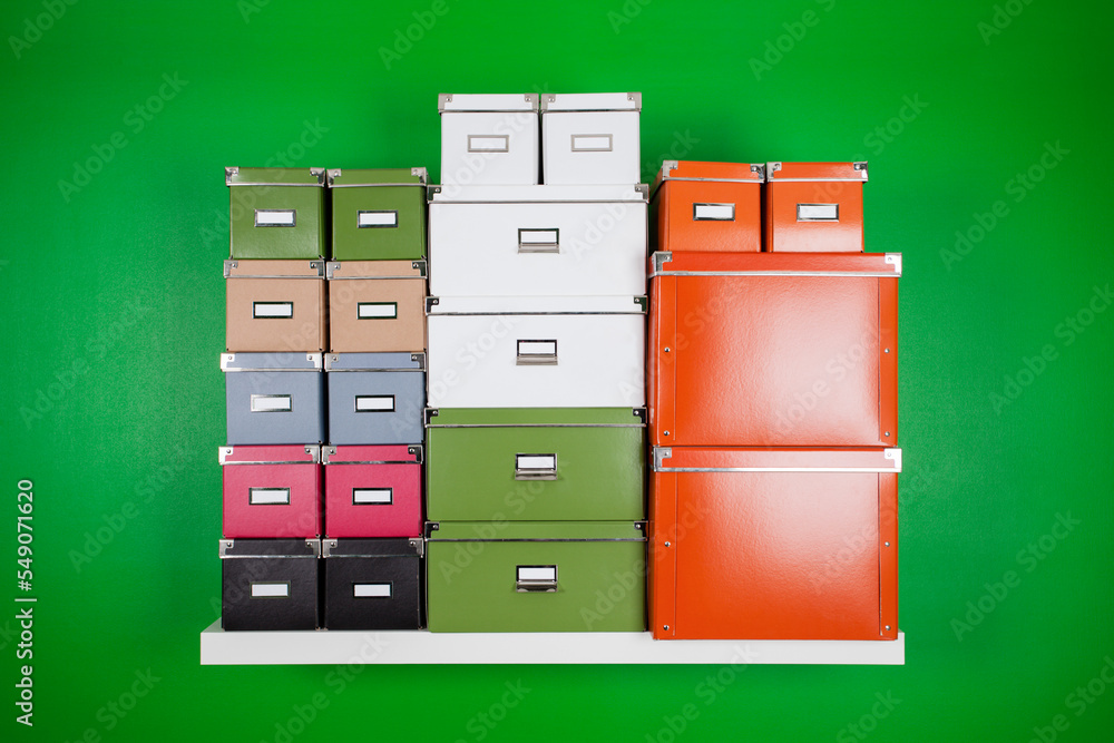 high stack of different color and size cardboard office boxes on a white bookshelf on a green wall. minimalistic composition on the theme of organization