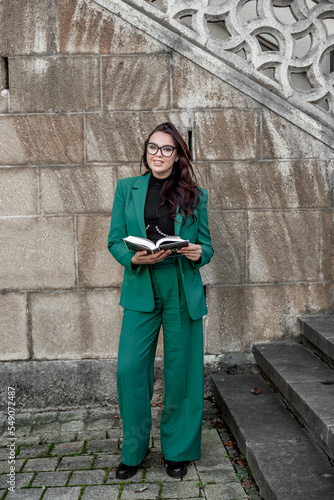 a girl in a green suit and glasses with a book, stands near the wall