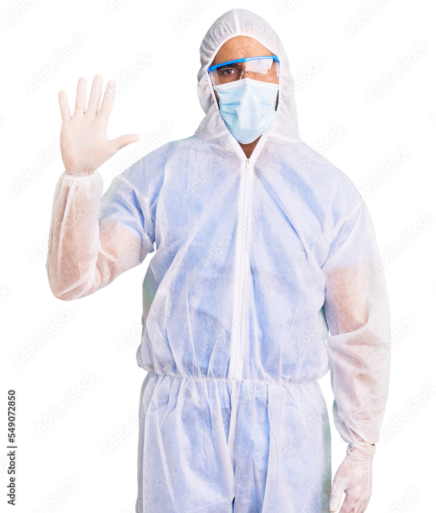 Young hispanic man wearing doctor protection coronavirus uniform and medical mask showing and pointing up with fingers number five while smiling confident and happy. Young hispanic man wearing doctor protection coronavirus uniform and medical mask showing and pointing up with fingers number five while smiling confident and happy.