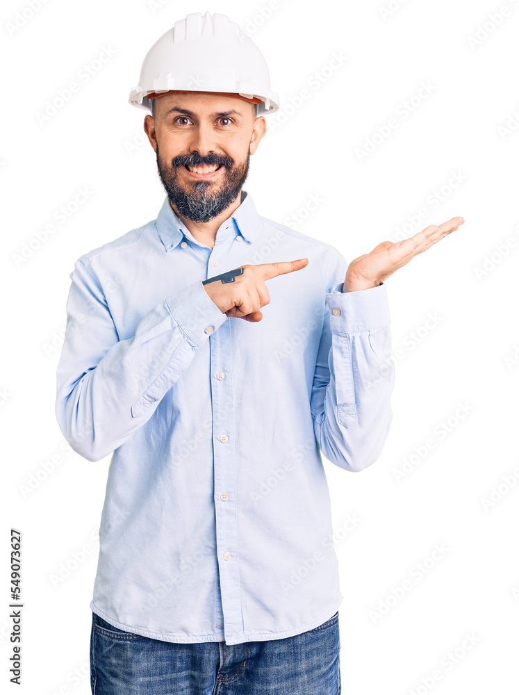 Young handsome man wearing architect hardhat amazed and smiling to the camera while presenting with hand and pointing with finger. Young handsome man wearing architect hardhat amazed and smiling to the camera while presenting with hand and pointing with finger.