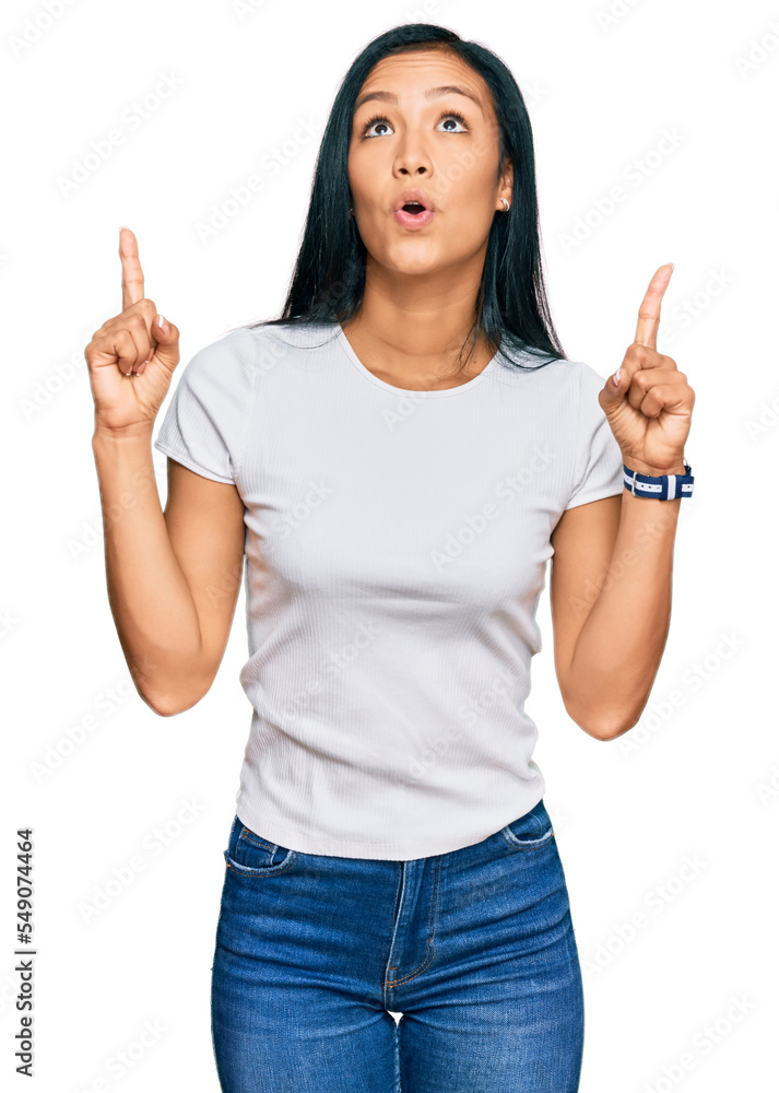 Beautiful hispanic woman wearing casual white tshirt amazed and surprised looking up and pointing with fingers and raised arms. Beautiful hispanic woman wearing casual white tshirt amazed and surprised looking up and pointing with fingers and raised arms.