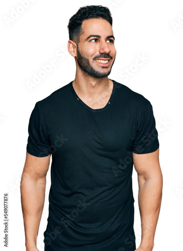 Hispanic man with beard wearing casual black t shirt looking away to side with smile on face, natural expression. laughing confident.